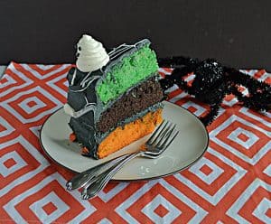 A close up of a slice of cake with a green, orange, and black layer, with two forks on the plate and a fake spider crawling onto the plate.