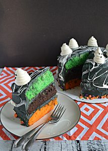 A slice of cake with a green, orange, and black layer, with two forks on the plate and the whole cake sitting behind it with the slice cut out.
