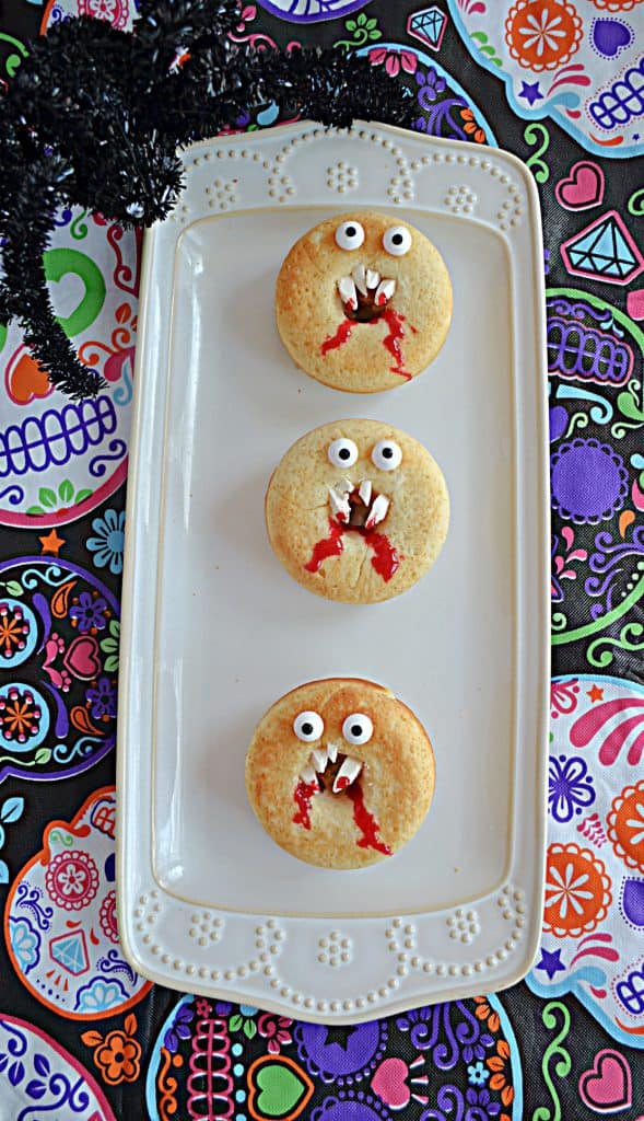 A platter with three vanilla donuts that look like vampire faces with fangs and edible eyes.