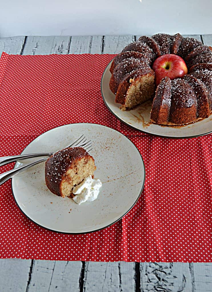 A side view of a plate with a slice of apple cider cake and whipped cream with two forks on the plate and the actual cake with a slice cut out and an apple in the middle in the background.