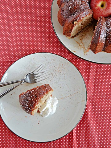 A top view of a plate with a slice of apple cider cake and whipped cream with two forks on the plate and the actual cake with a slice cut out in the background.