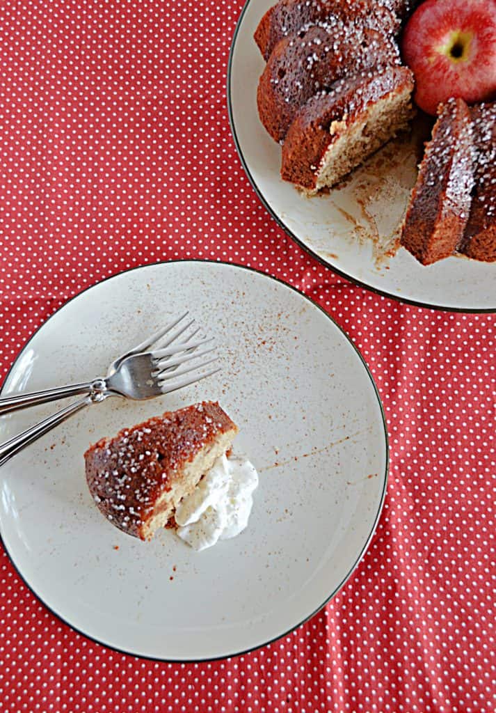 A top view of a plate with a slice of apple cider cake and whipped cream with two forks on the plate and the actual cake with a slice cut out in the background.