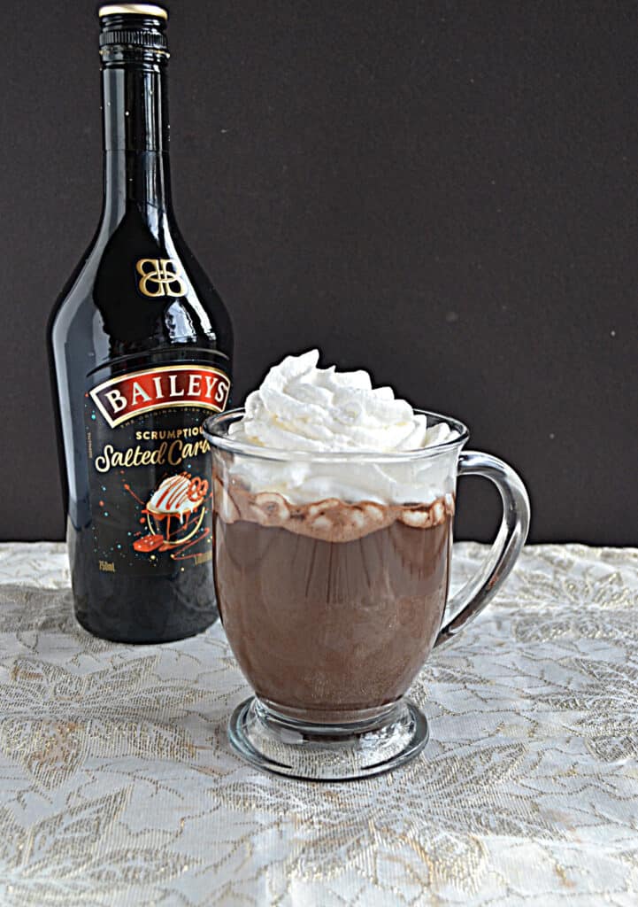 A mug of hot chocolate topped with whipped cream wwith a bottle of Bailey's in the background.