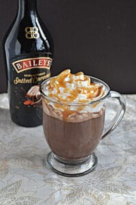 A mug of hot chocolate topped with whipped cream and caramel sauce with a bottle of Bailey's in the background.