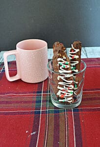 A pink coffee mug next to a glass with three Chocolate Biscotti drizzled with white chocolate