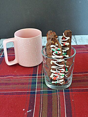A pink coffee mug next to a glass with three Chocolate Biscotti drizzled with white chocolate