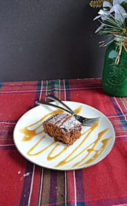 A plate with a slice of gingerbread cake drizzled with caramel sauce and two forks on the place with a green vase behind the plate.