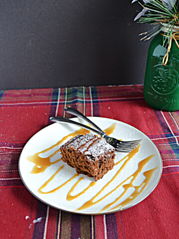 A plate with a slice of gingerbread cake drizzled with caramel sauce and two forks on the place with a green vase behind the plate.