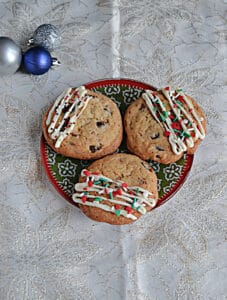 A plate of 3 cookies drizzled with white chocolate and sprinkles with a pile of ornaments in the background.