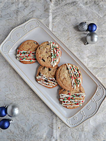 A white platter piled with cookies drizzled in white chocolate with sprinkles and ornaments on both sides of the platter.