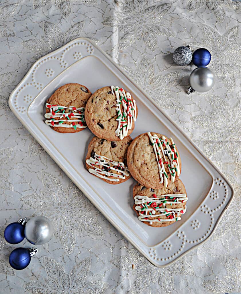 A white platter piled with cookies drizzled in white chocolate with sprinkles and ornaments on both sides of the platter. 
