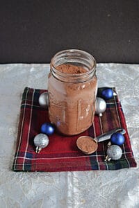 A jar of hot chocolate mix with a spoon of hot chocolate on the side along with blue and silver ornaments.
