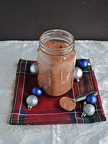 A jar of hot chocolate mix with a spoon of hot chocolate on the side along with blue and silver ornaments.