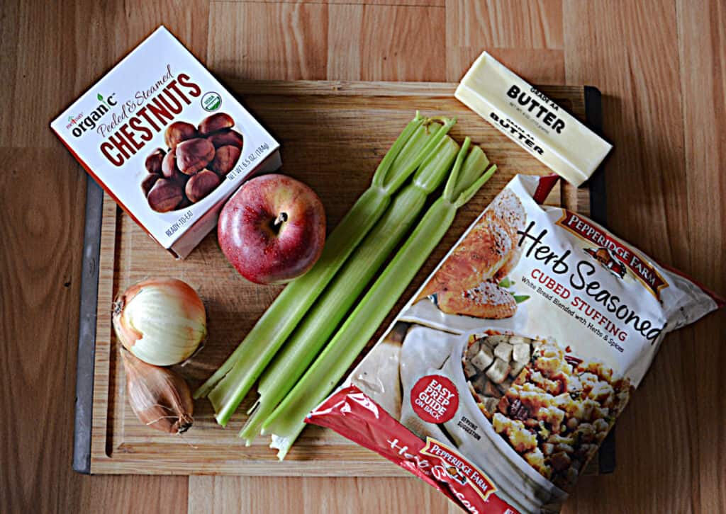 A cutting board with a box of chestnuts, an apple, and onion, three stalks of celery, a stick of butter, and a bag of bread cubes on it. 