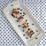 A white platter turned at an angle with squares of chocolate chip cookie dough fudge with chocolate chips sprinkled on top.