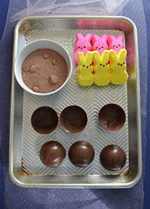 A cookie sheet with a cup of hot cocoa mix, six bunny marshmallows, and six half spheres of chocolate.