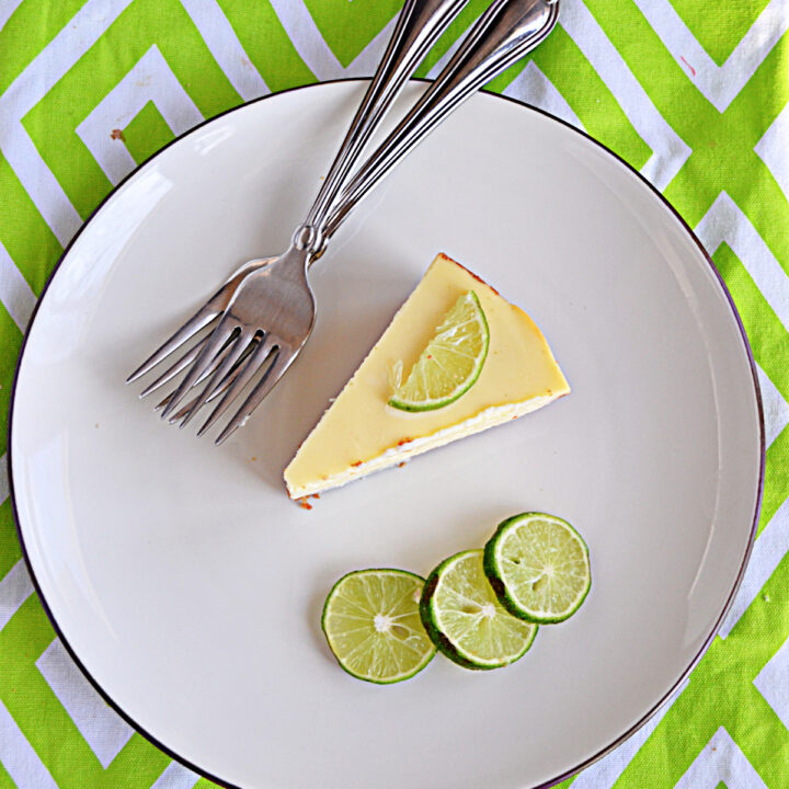 A plate with a slice of cheesecake, three slices of lime, and two forks on the plate.