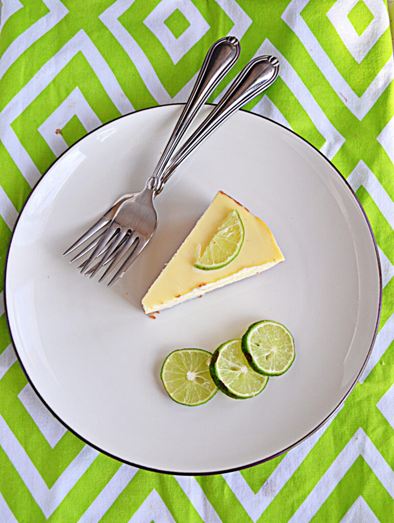 A plate with a slice of cheesecake, three slices of lime, and two forks on the plate.