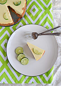 A plate with a slice of cheesecake, three slices of lime, and two forks on the plate and the entire cheesecake behind the plate.
