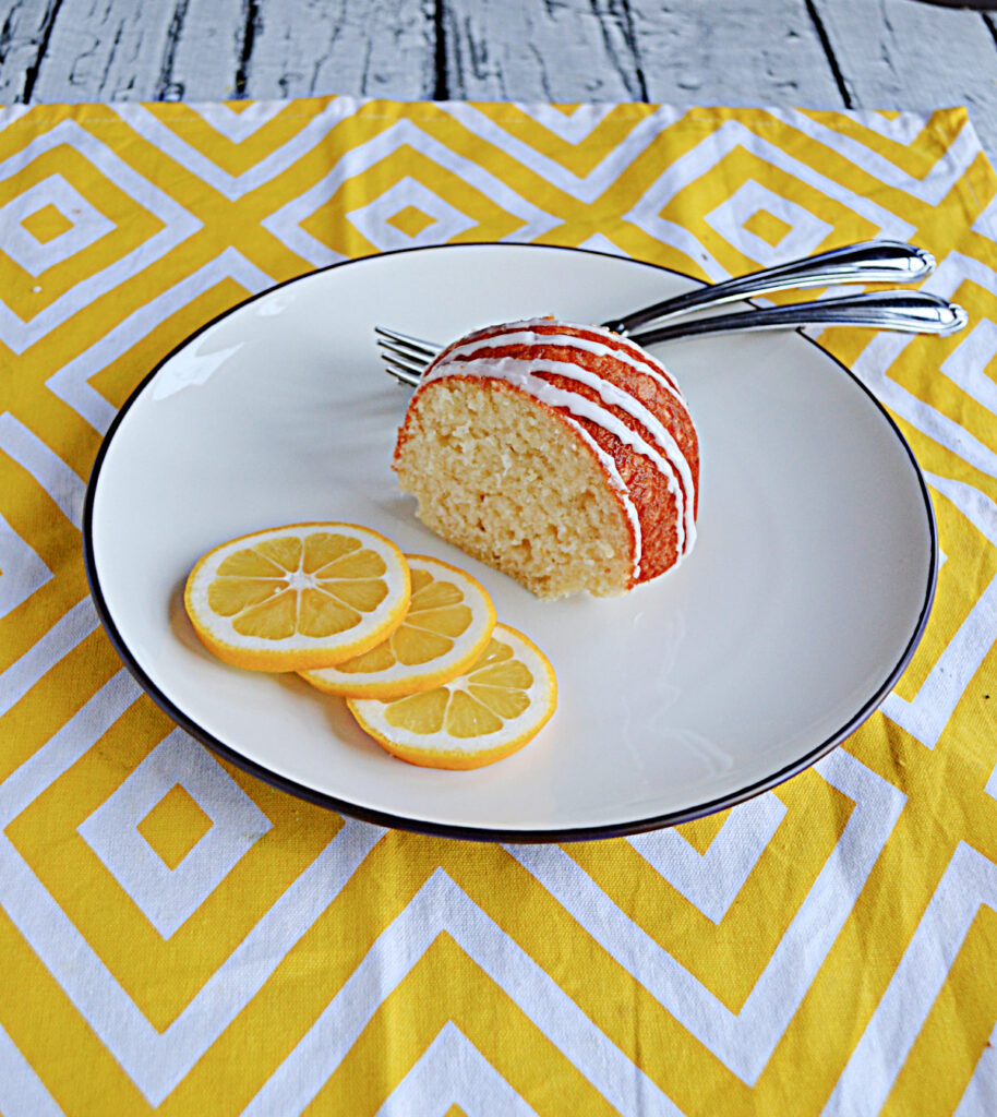 A slice of Bundt cake on a plate with a white glaze drizzle on it, two forks behind the cake, and three lemon slices in front of the cake.