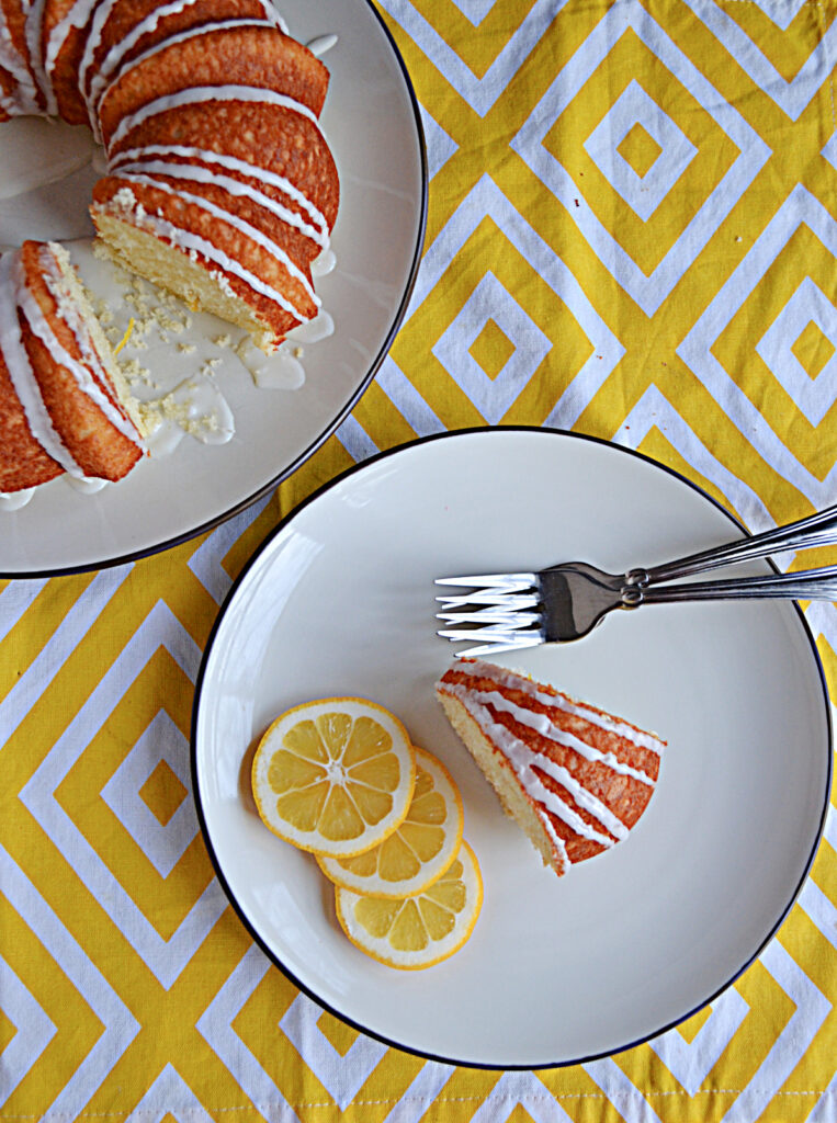 A top view of a plate with a slice of Bundt cake, 3 slices of Meyer lemon, and two forks with the entire Bundt cake behind it and off to the side. 