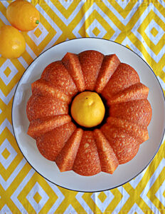 A plate with a Bundt cake on it with a lemon in the center of the cake and lemons off to the side behind the cake.