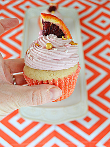 A close up view of a Blood Orange Creamsicle Cupcake with a wedge of blood orange on top and a hand holding the cupcake.