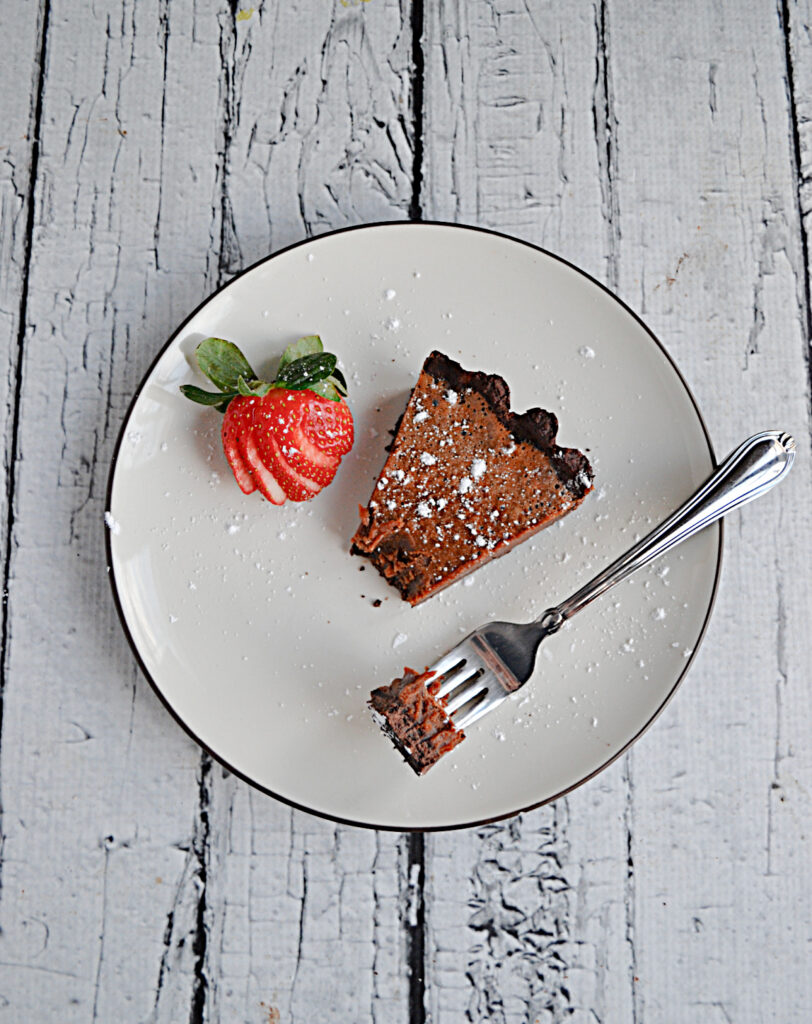 A plate with a slice of Chocolate Caramel Tart with a bite taken out of it, a fork with a bite of tart on it, and a strawberry on it.