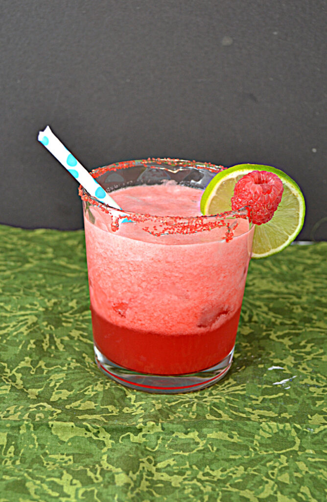 A glass of deep pink Raspberry Margaaritas with a straw in it and a lime slice and a raspberry on the rim. 