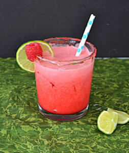 A glass of Raspberry Margarita with a straw in it and a lime slice and a raspberry on the rim.