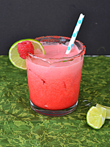 A glass of Raspberry Margarita with a straw in it and a lime slice and a raspberry on the rim.
