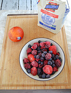 A cutting board with a bag of sugaar, a bowl of frozen berries, and an orange.