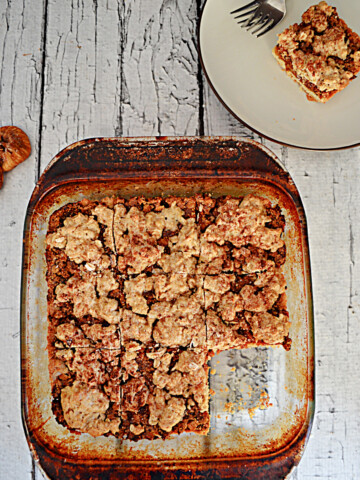 A baking dish with fig crumble bars in them with one piece missing and a plate with a crumble bar on it with 2 forks.