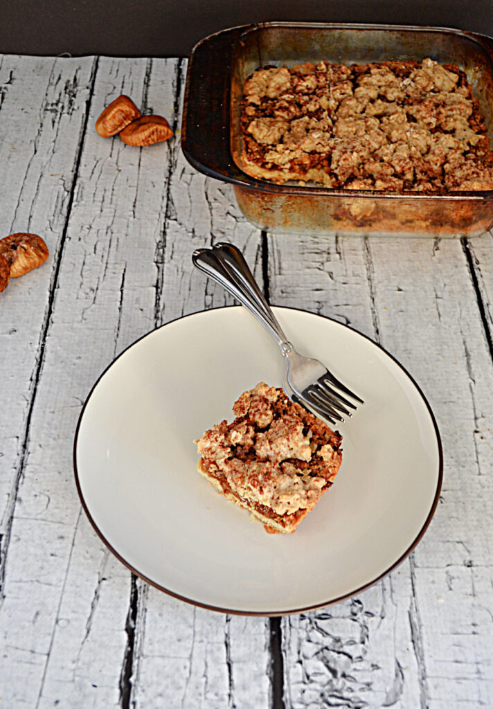 A close up view of a plate with a fig crumble bar and 2 forks on it and a baking dish behind it with a pan of fig bars in it. 