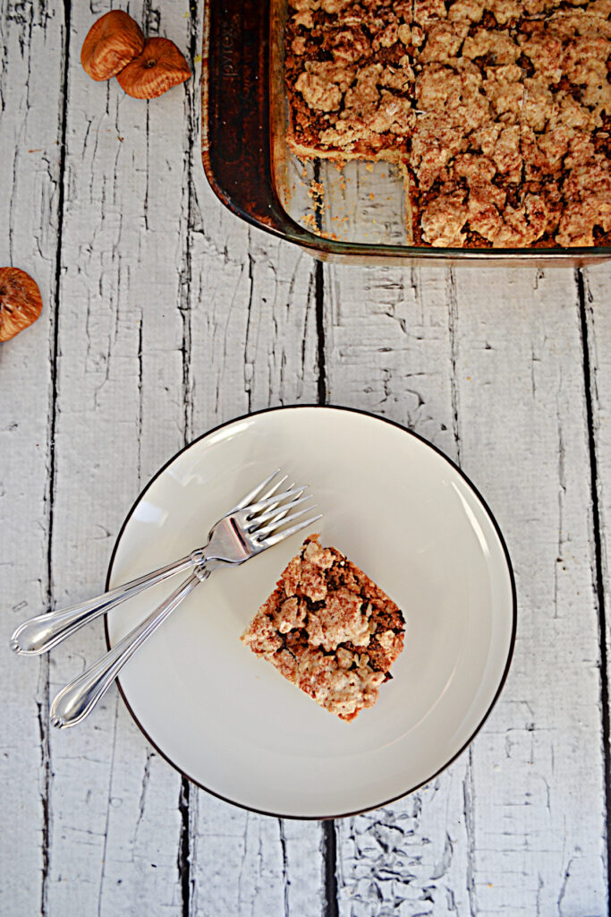 A plate with a fig crumble bar and 2 forks on it and a baking dish behind it with a pan of fig bars in it. 