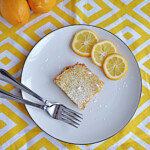 A plate with a lemon bar, three slices of lemon, and two forks on a plate with 2 Meyer lemons behind the plate.