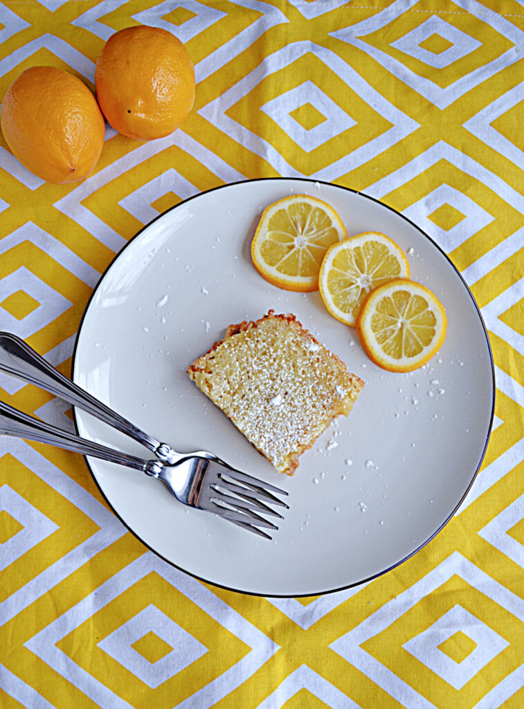 A plate with a lemon bar, three slices of lemon, and two forks on a plate with 2 Meyer lemons behind the plate.