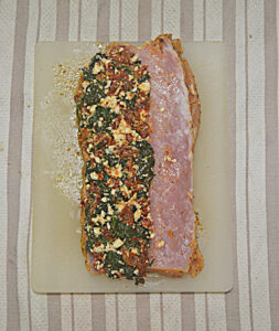 A cutting board with a pork loin split in half and spread with spinach, sun dried tomatoes, and feta.