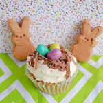 A cupcake with a chocolate bird's nest and candy eggs on top and two Peeps Bunnies behind the cupcake.