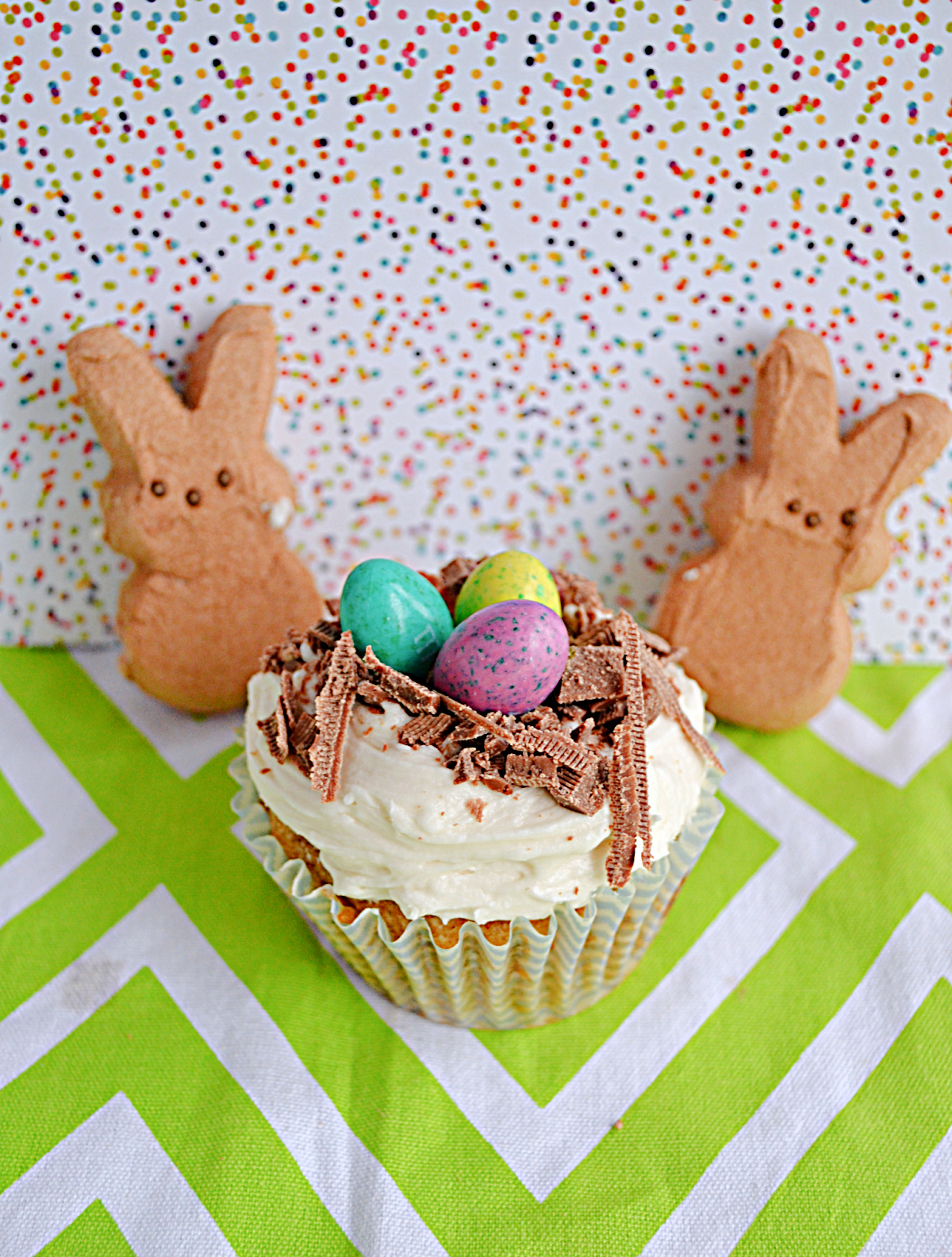 A cupcake with a chocolate bird's nest and candy eggs on top and two Peeps Bunnies behind the cupcake.