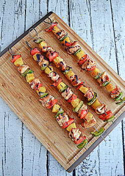 A cutting board with four grilled chicken, bell pepper, and pineapple kabobs on it.