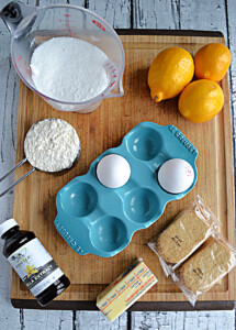 A cutting board with three Meyer lemons, two eggs, a cup of gluten free flour, a cup of sugar, a bottle of vanilla, a stick of butter, and two packs of oat grahams on it.
