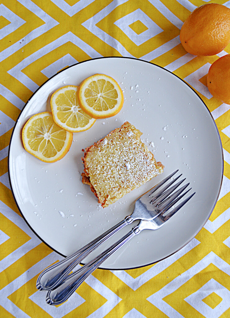 A top view of a plate with a lemon bar square, three slices of lemon, and two forks on it with a lemon behind the plate.