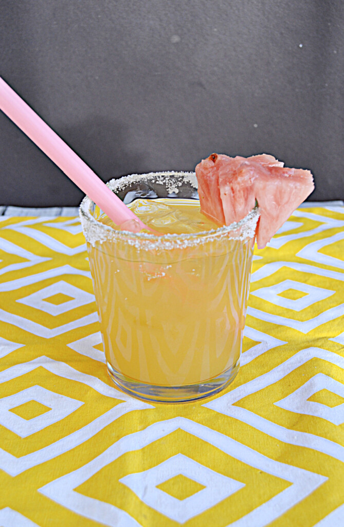 A yellow cocktail with a pink straw, sugar rim, and pink pineapple garnish.