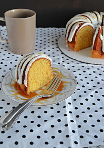 A close up of a slice of Bundt Cake with two forks on the plate, a mug in the background, and a Bundt Cake in the background.