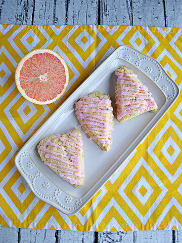 A white platter of three grapefruit cardamom scones with pink glaze on top and half a grapefruit behind the platter.