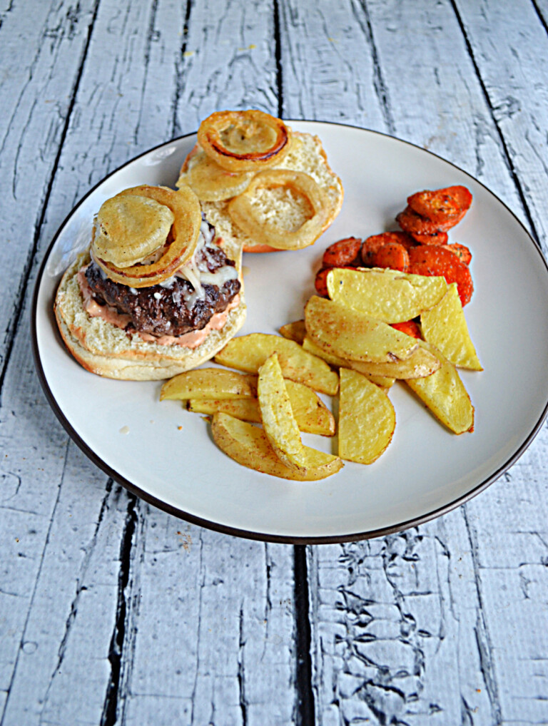 A hamburger topped with fried onions and pickles, a side of potato wedges, and carrots.