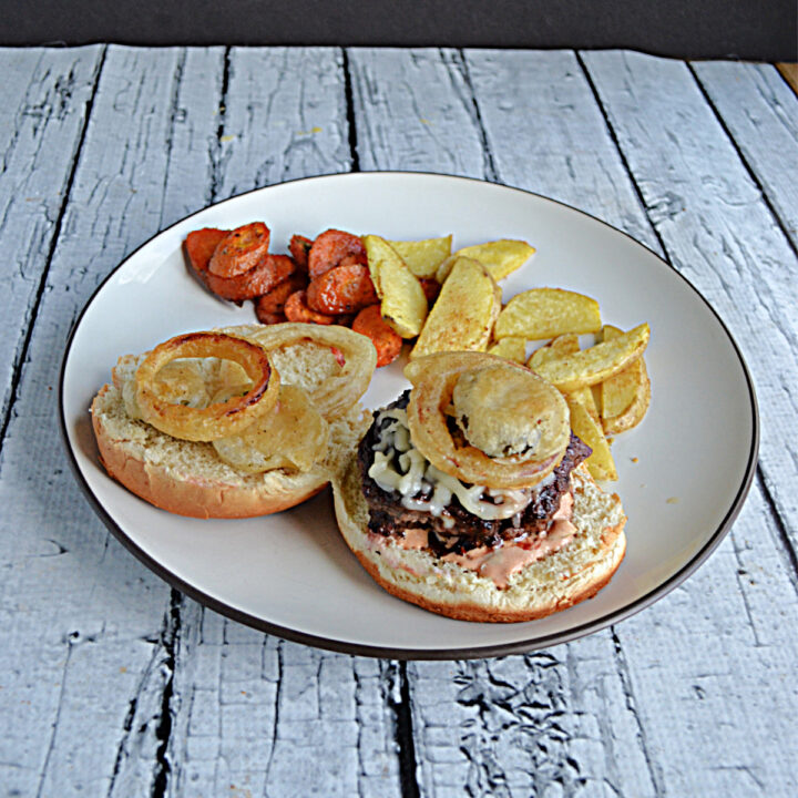 A plate with a burger topped with fried pickles, onions, and burger sauce with potato wedges and carrots.
