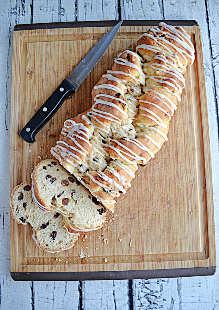 A cutting board with a golden brown loaf of Rum Raisin Bread with a glaze drizzled on top and two slices cut off them bread. 