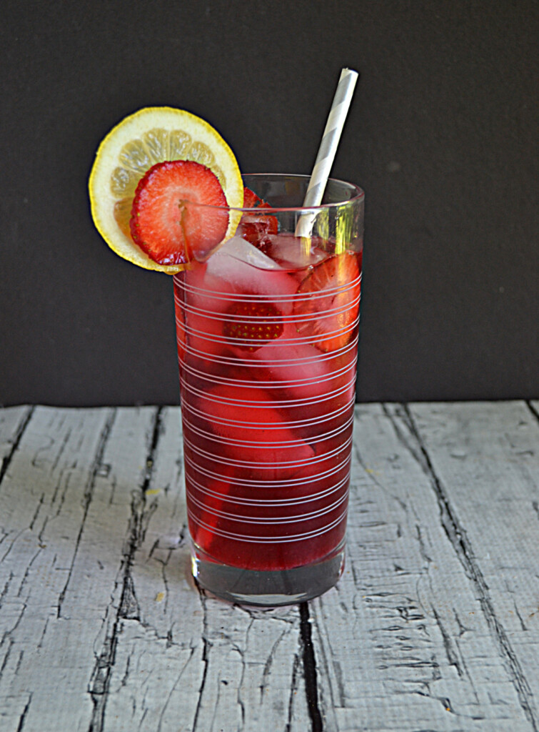 A glass of berry iced tea with a lemon slice and strawberry slice and a straw in the glass.